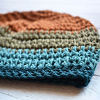 Close-up of a crocheted dbeanie in orange, green, and teal blue colors on a white wooden surface.