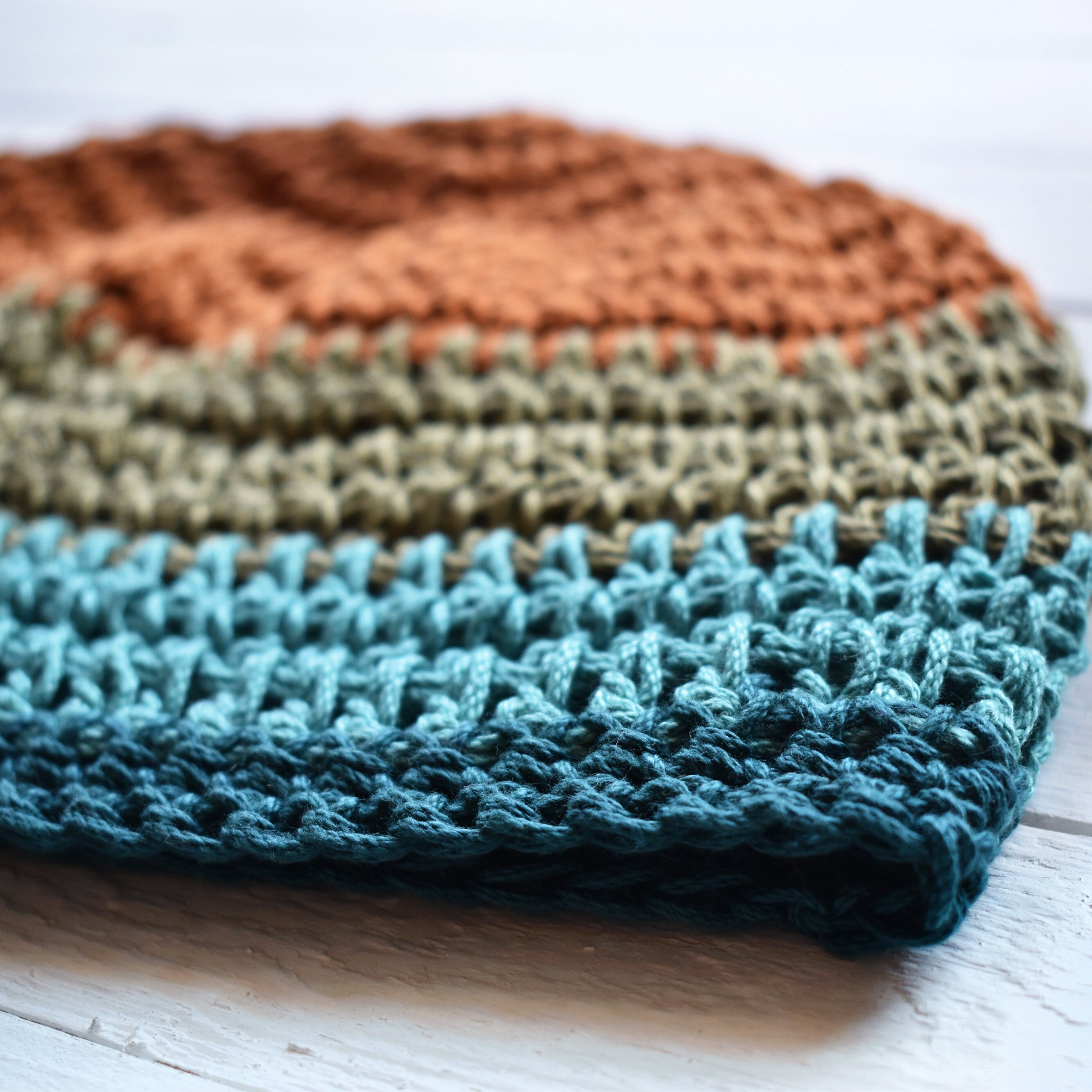 Close-up of a crocheted dbeanie in orange, green, and teal blue colors on a white wooden surface.