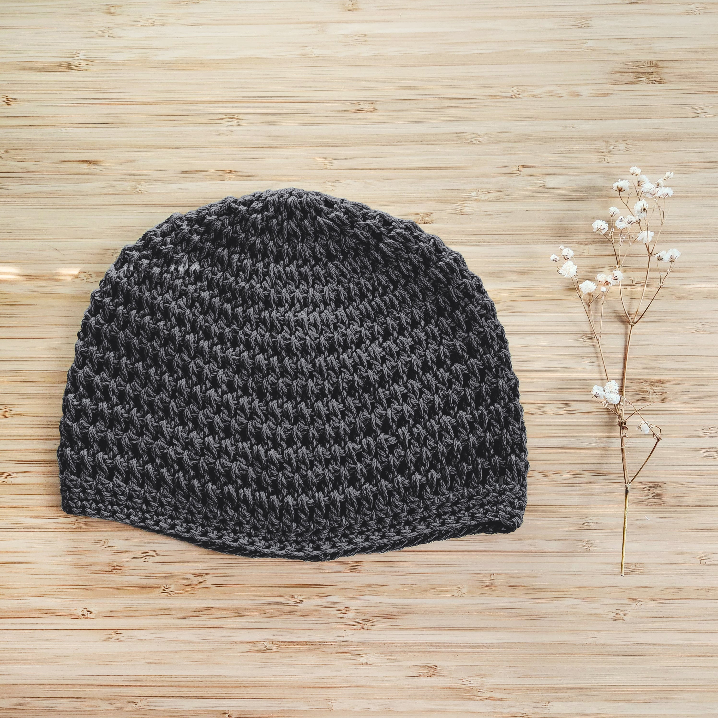 Dark gray crocheted beanie on a wooden surface next to a sprig of dried flowers.