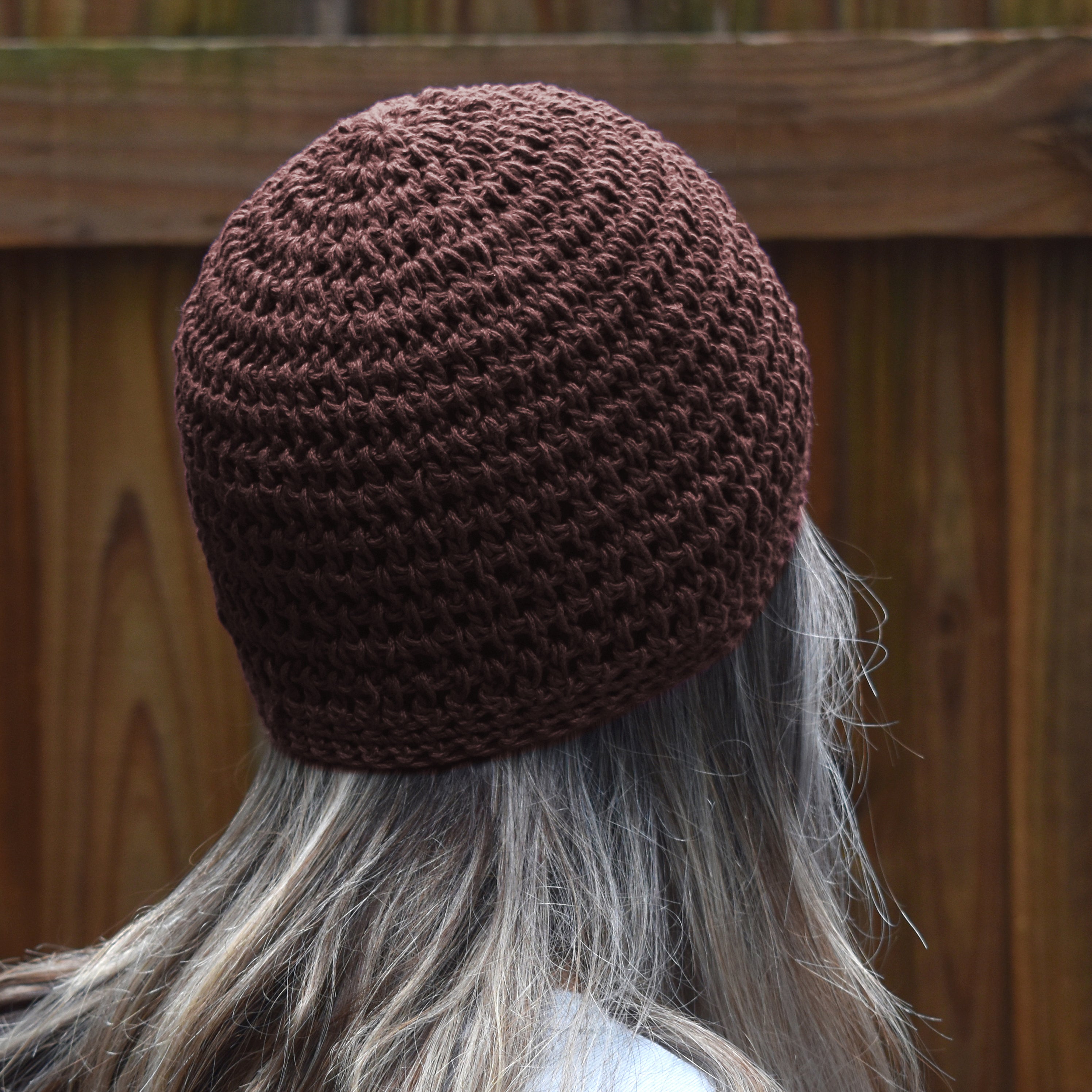 Brown knitted beanie worn by a person with gray hair, against a wooden fence background.