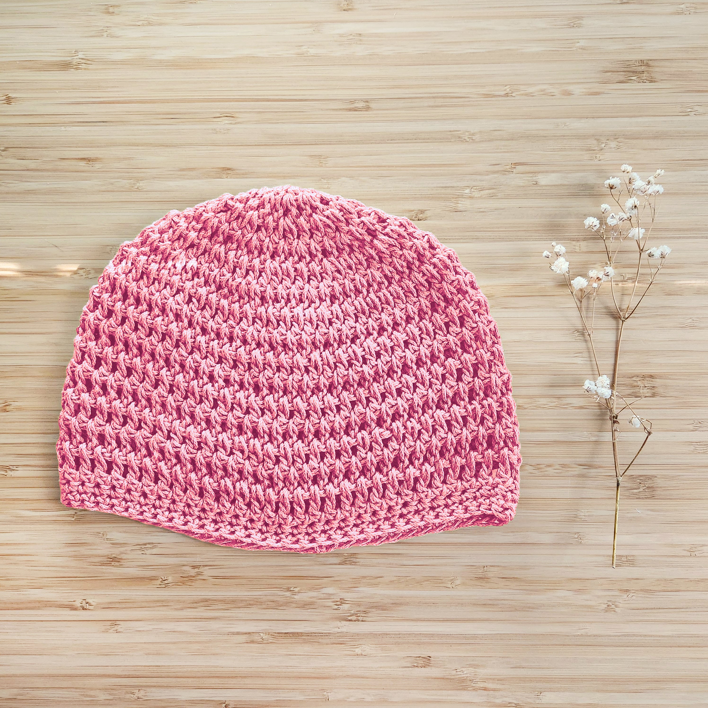 Pink crocheted beanie on a wooden surface with a small branch of flowers.