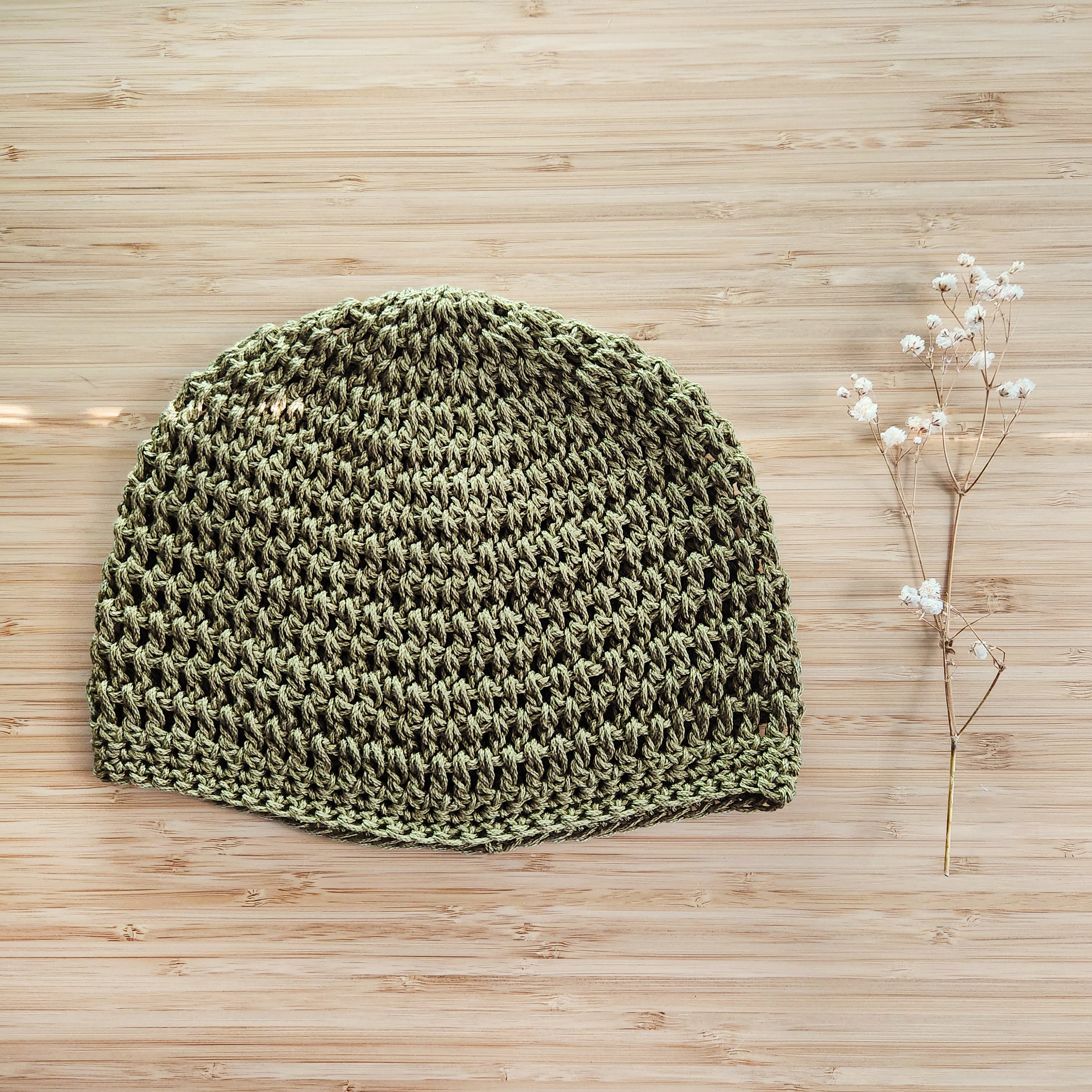 Green crocheted beanie on a wooden surface with a sprig of flowers.