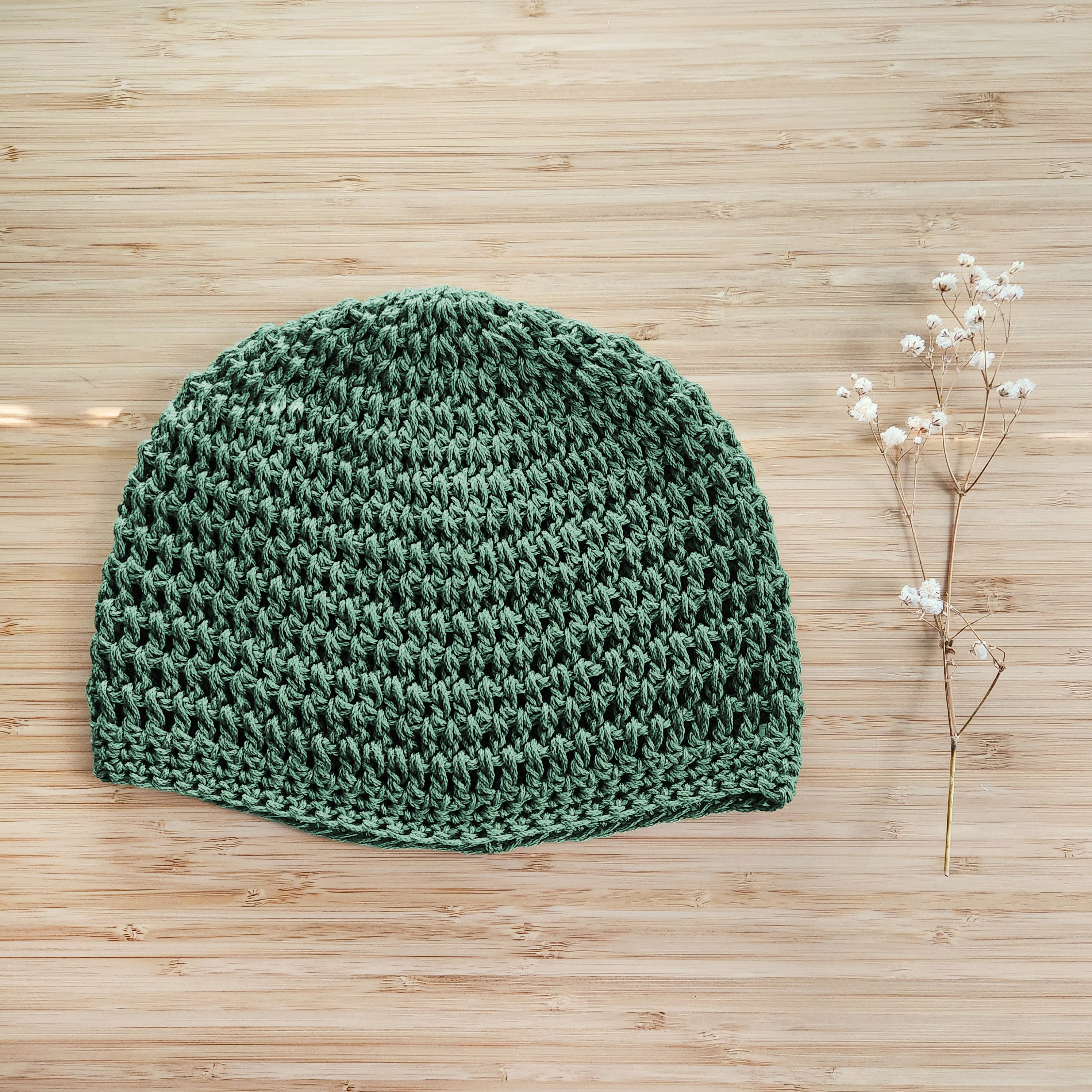 Green crocheted beanie on a wooden surface with a sprig of dried flowers.