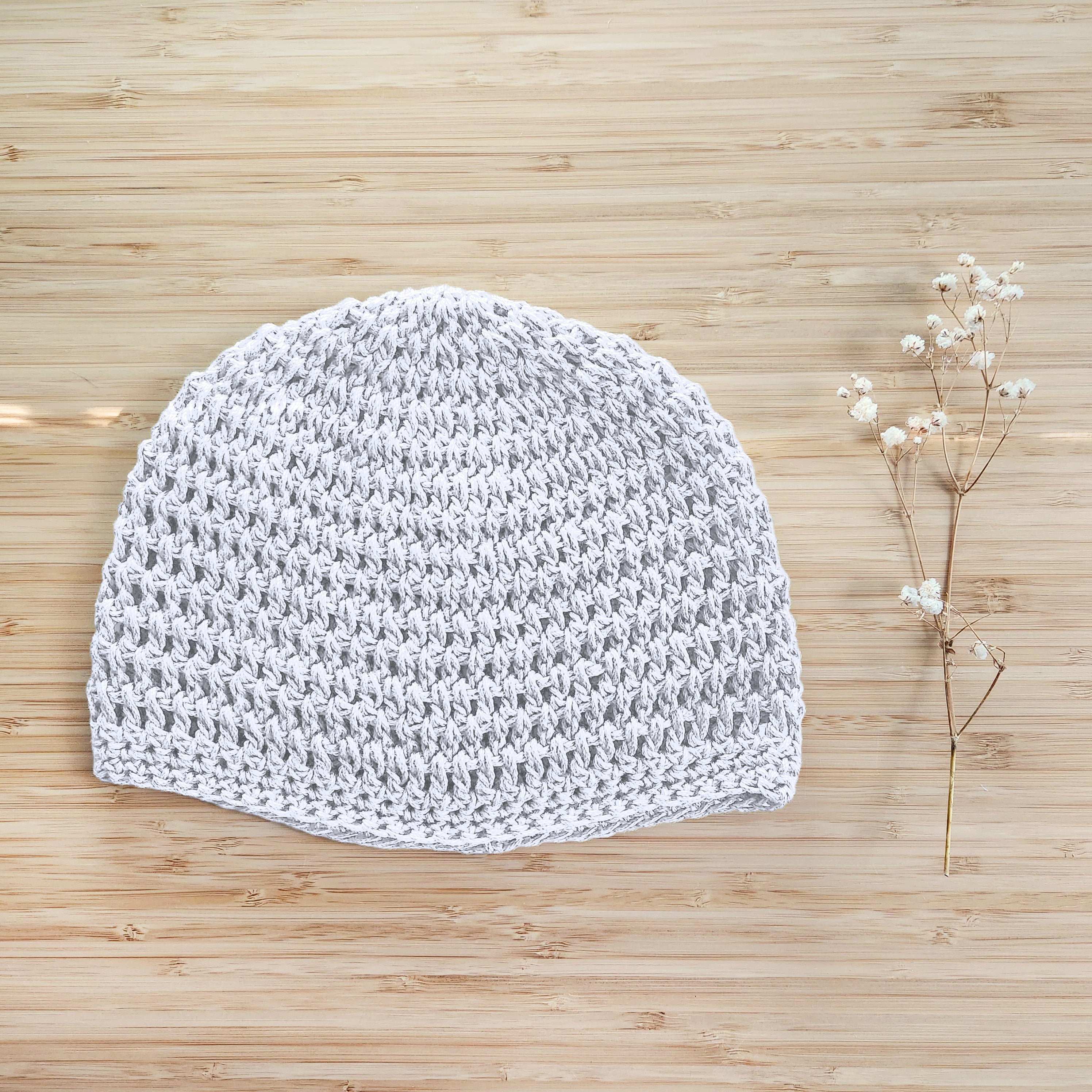 White crocheted beanie on a wooden surface with a small branch of flowers.