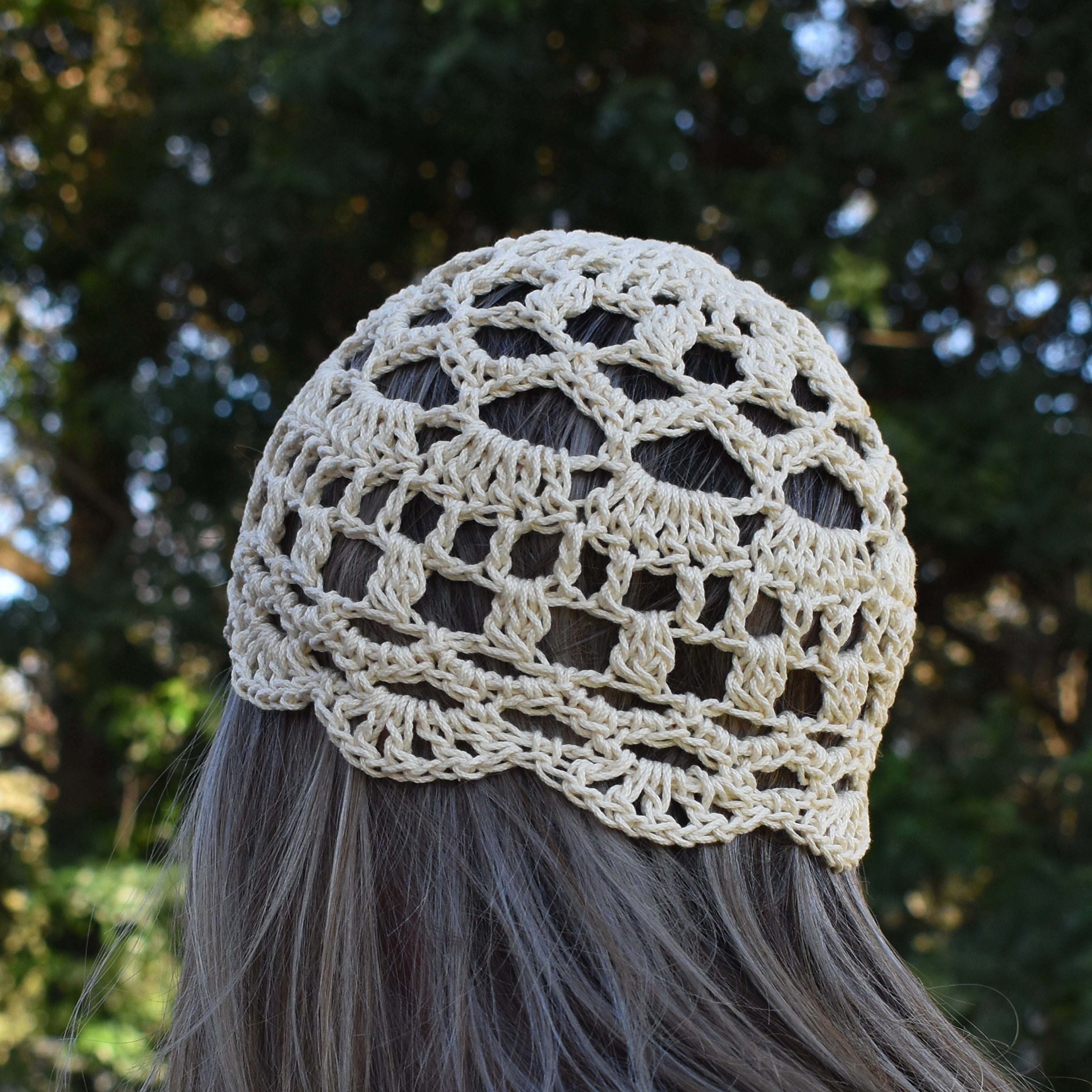 Beige crochet lace skull cap worn by a person with blurred green foliage in the background.