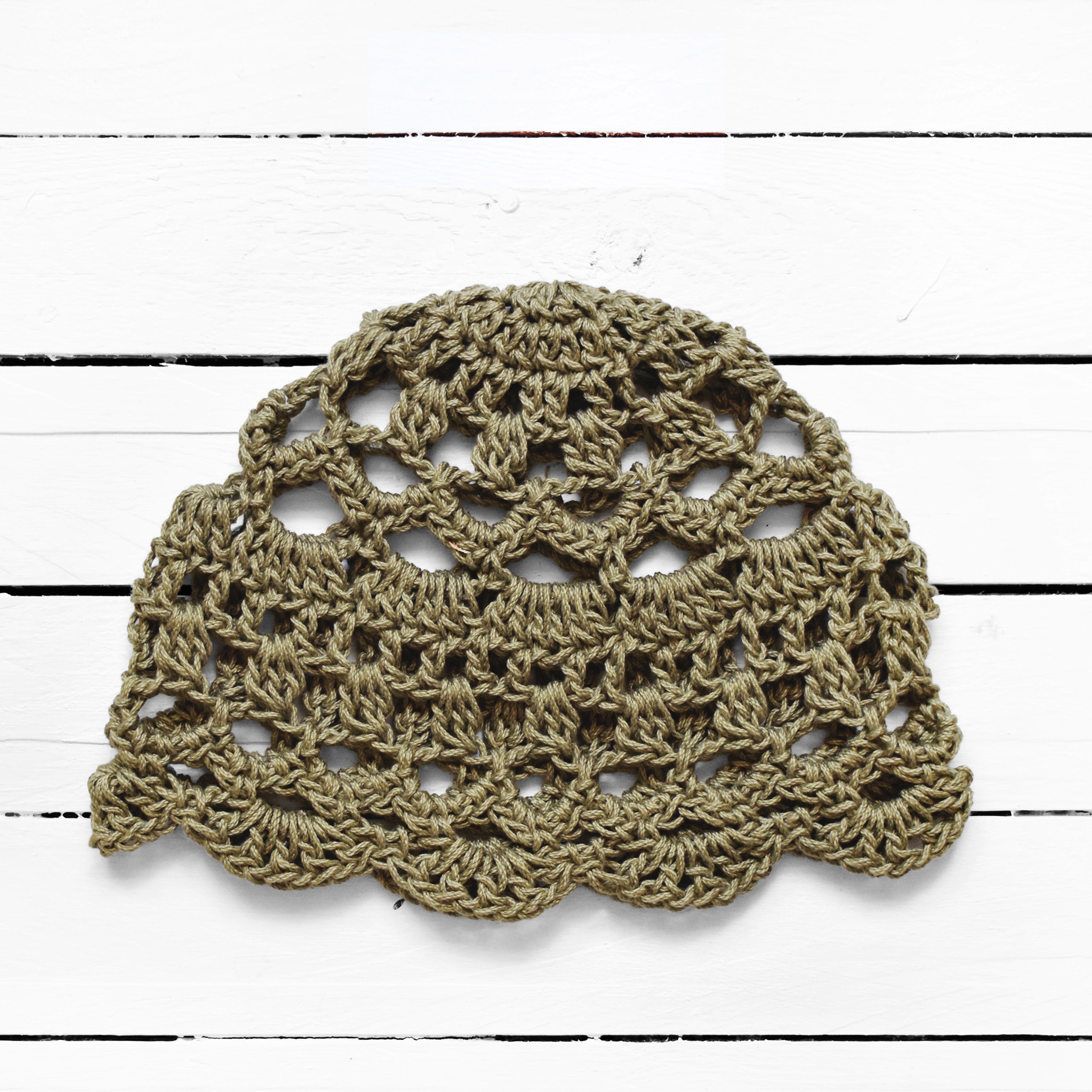 Sage green crocheted lace cap on a white wooden surface.