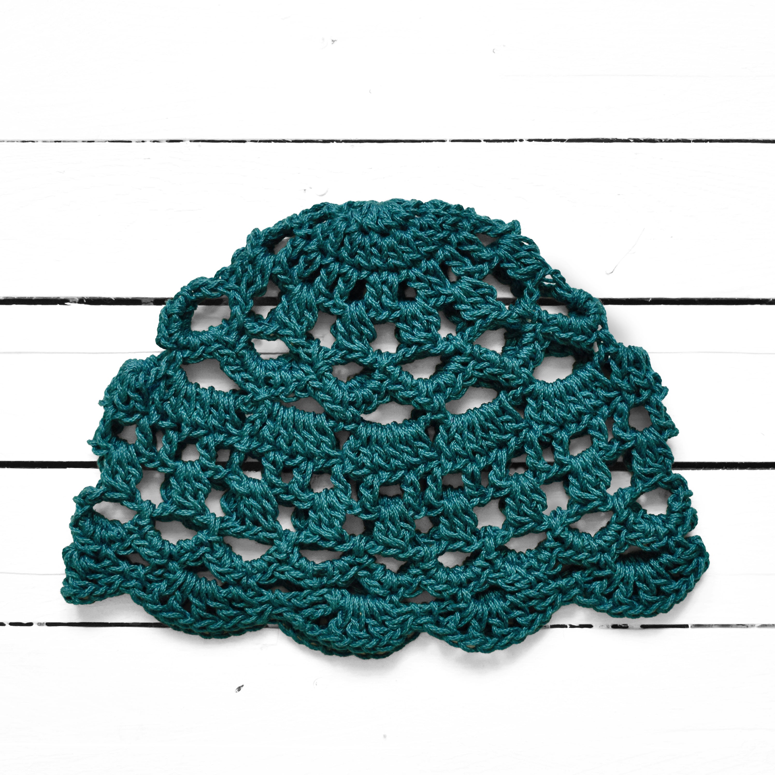 Dark teal crocheted lace summer cap on a white wooden surface.