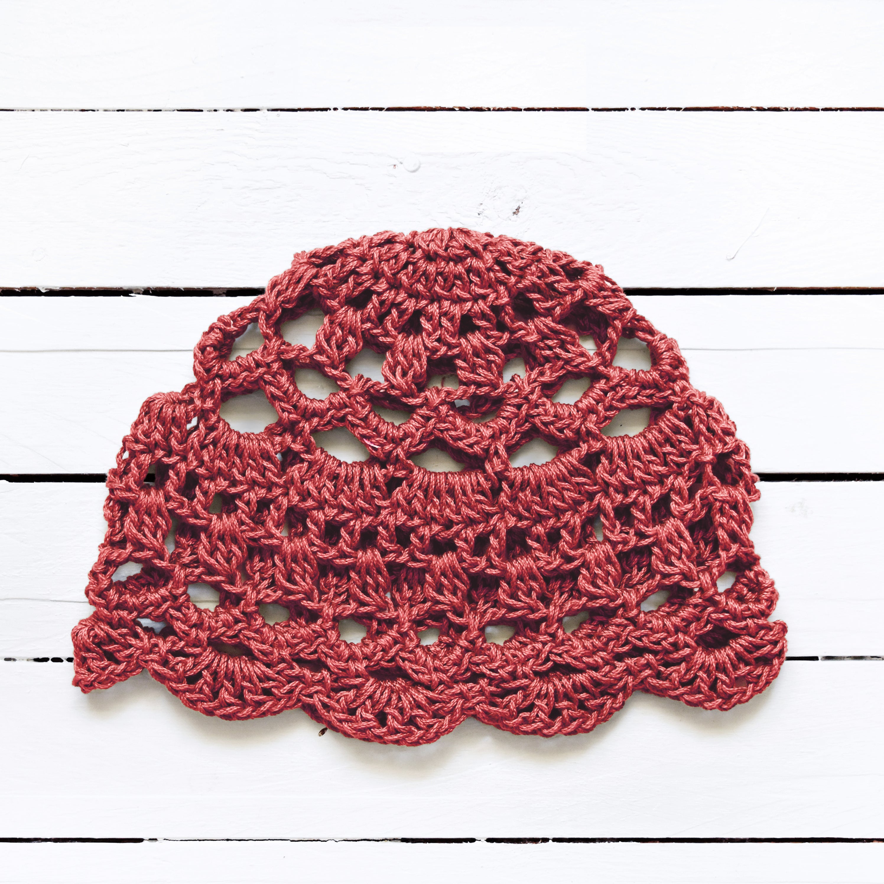 Boho crocheted lace cap in an earthy terracotta pink color on a white wooden surface.