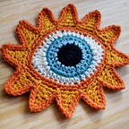 Close-up view of a crocheted sun-shaped motif with a blue eye in the center on a wooden surface.