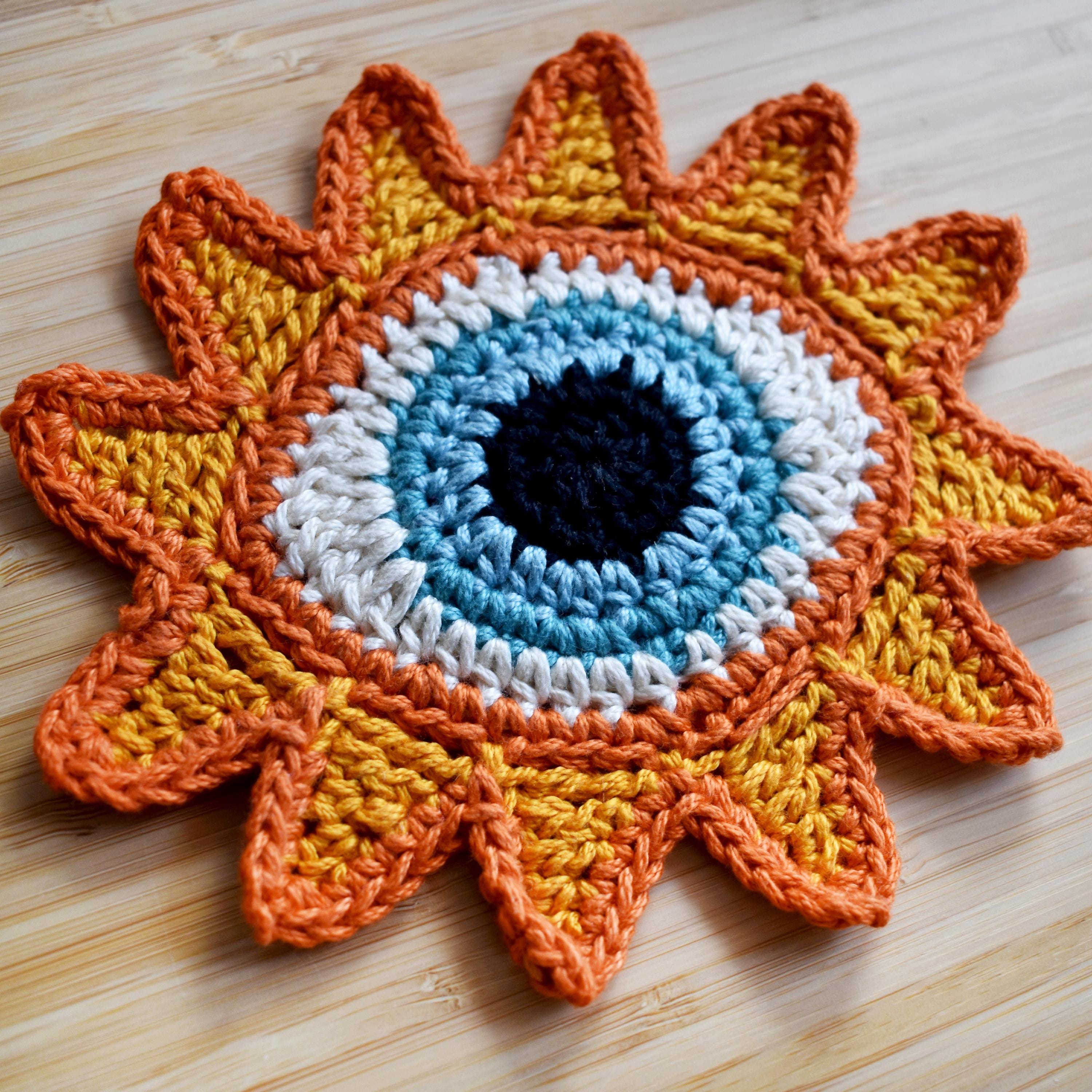 Close-up view of a crocheted sun-shaped motif with a blue eye in the center on a wooden surface.