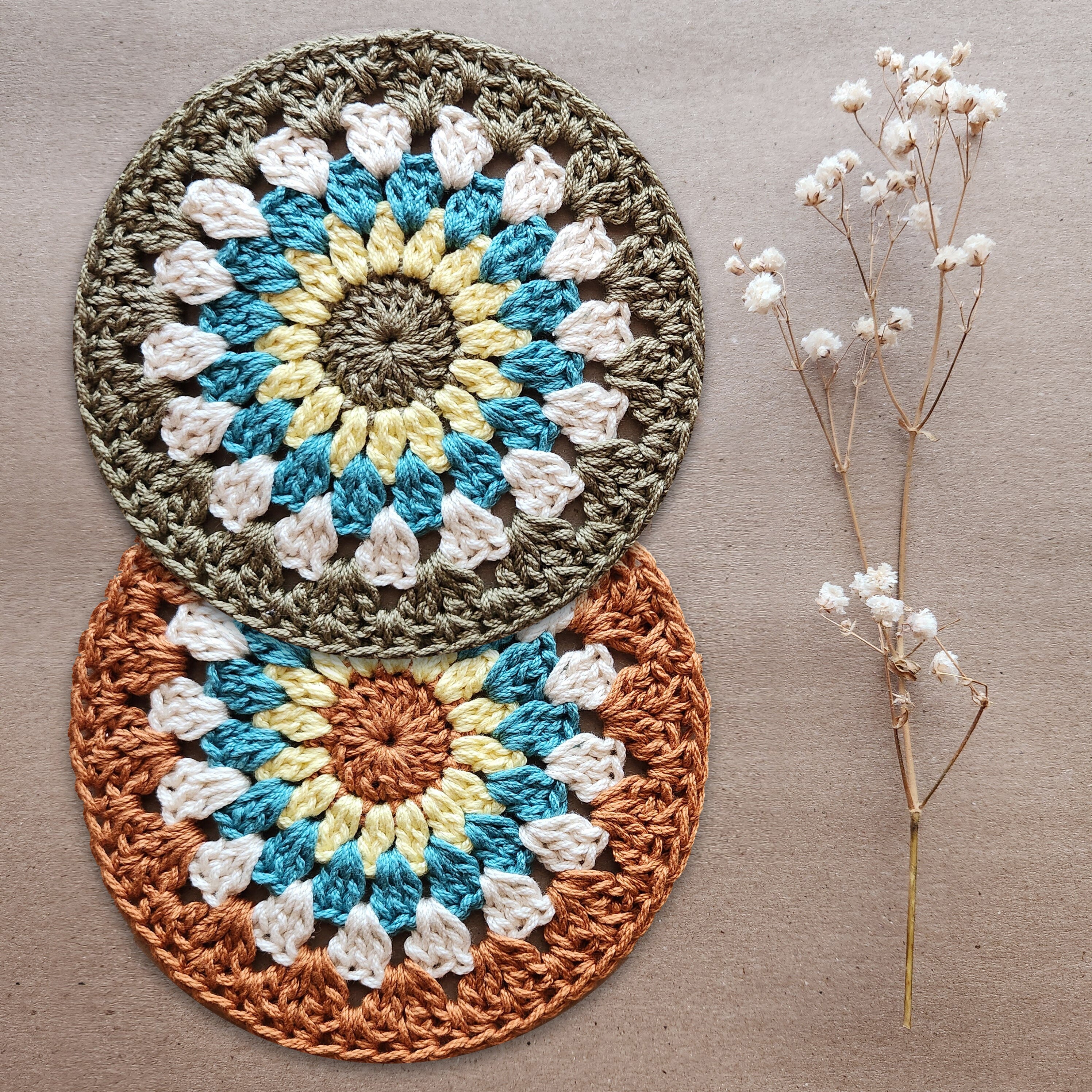 Two crocheted coasters with a floral pattern on a natural kraft background, accompanied by a dried sprig of flowers.