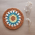 A larger crocheted mandala coaster with brown, blue, and white colors next to a sprig of flowers on a natural kraft background.