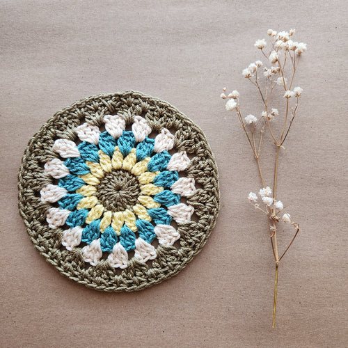 Large round crocheted coaster with a green, blue, yellow, and beige mandala pattern next to a sprig of flowers on a natural kraft background.