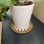 White ceramic pot with a plant on a textured surface, featuring a decorative woven coaster.