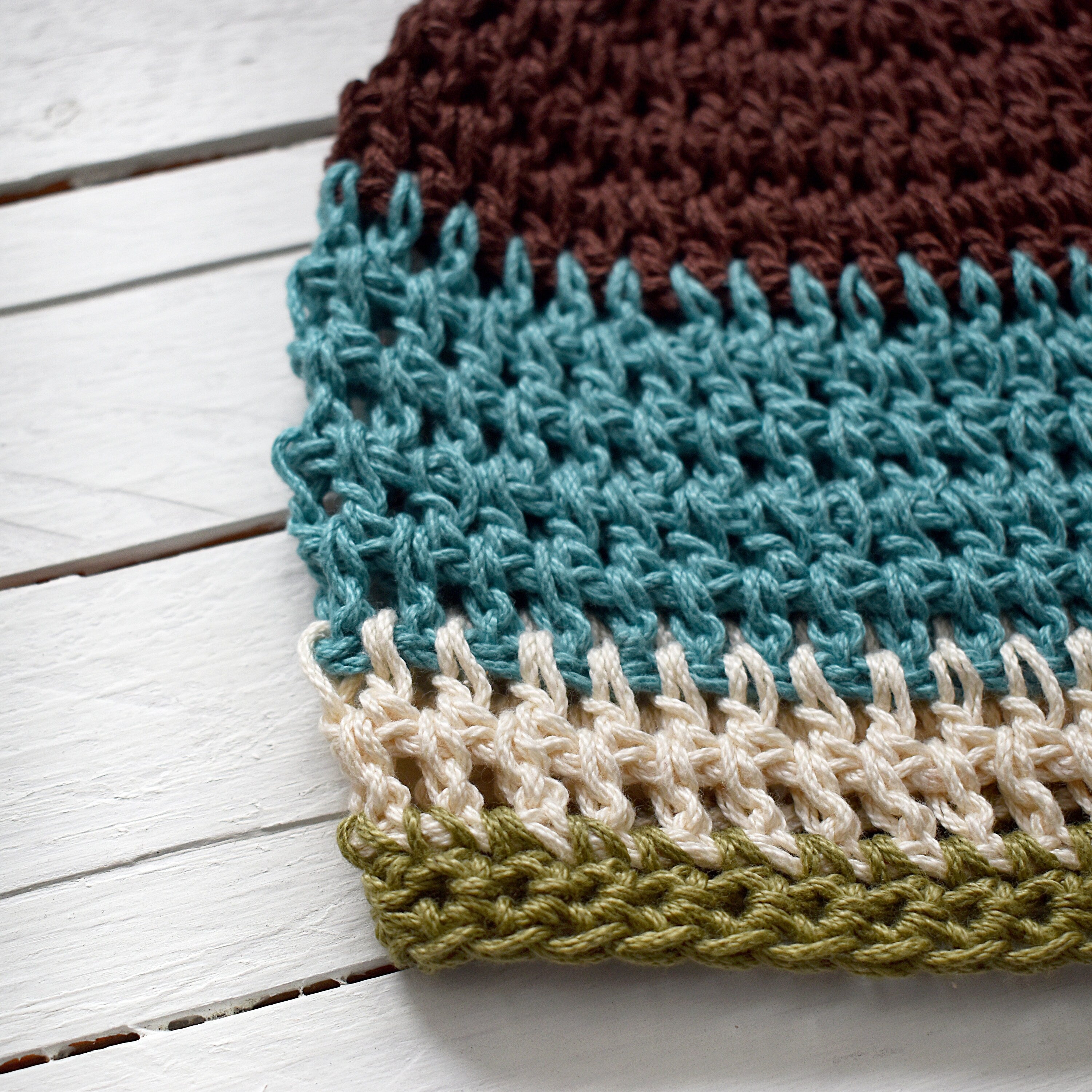 Close-up of a crocheted hat with brown, blue, beige, and green colors on a white wooden surface.
