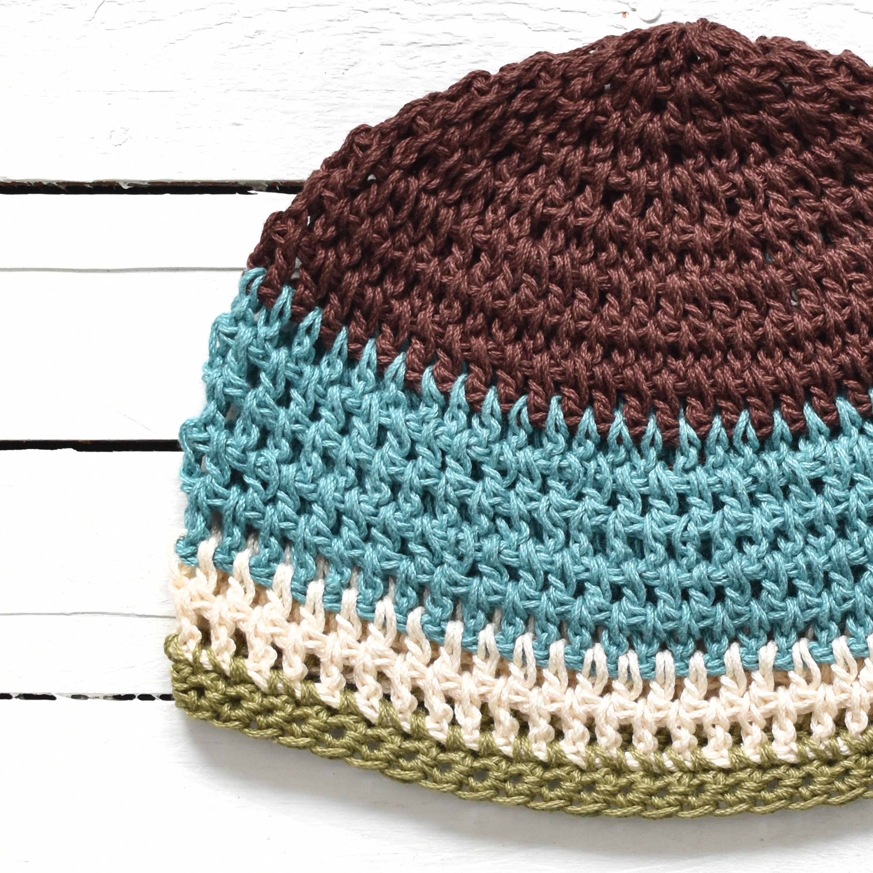Crocheted beanie with brown, blue, beige, and green colors on a white wooden surface.