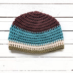 Crocheted beanie with brown, blue, beige, and green colors on a white wooden surface.