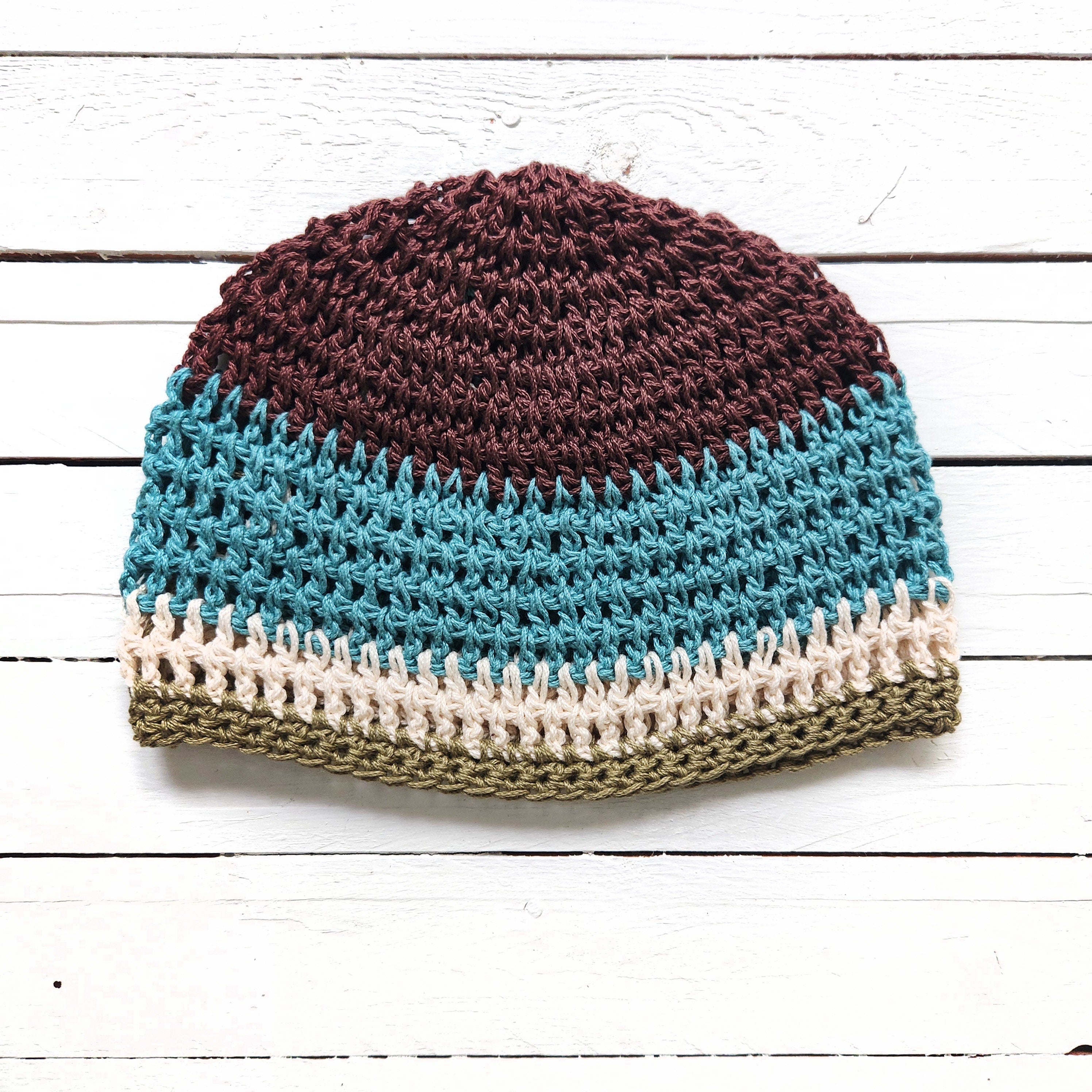 Crocheted beanie with brown, blue, beige, and green colors on a white wooden surface.