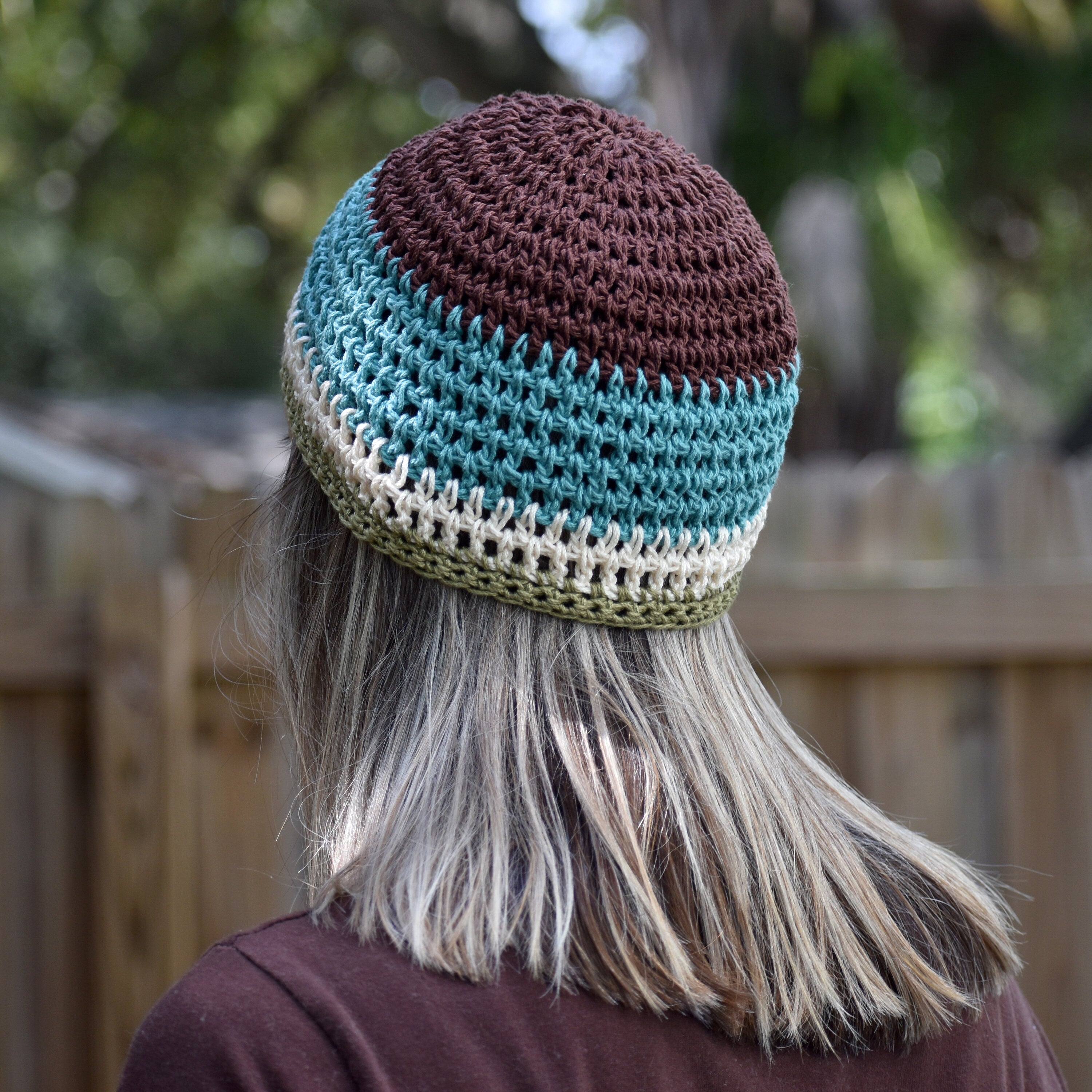 Person wearing a crocheted beanie with brown and blue stripes against a blurred outdoor background