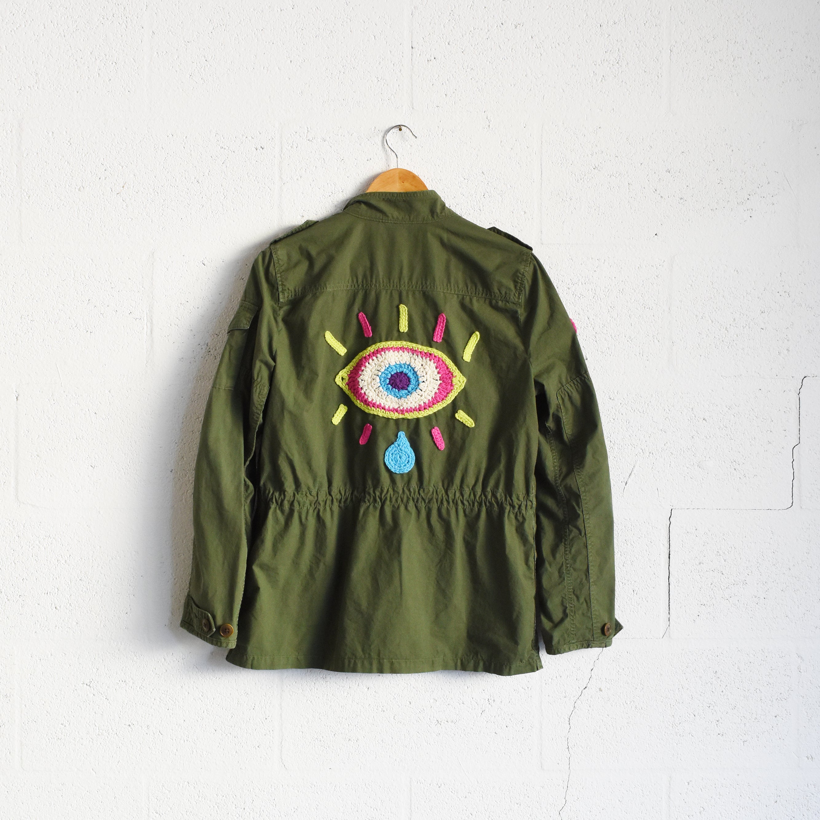 Upcycled army green utility jacket with colorful crochet eye design hanging on a white cement background wall.