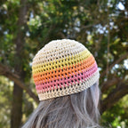 A person wearing a beige crochet beanie with yellow, orange, and pink stripes outdoors with trees in the background.
