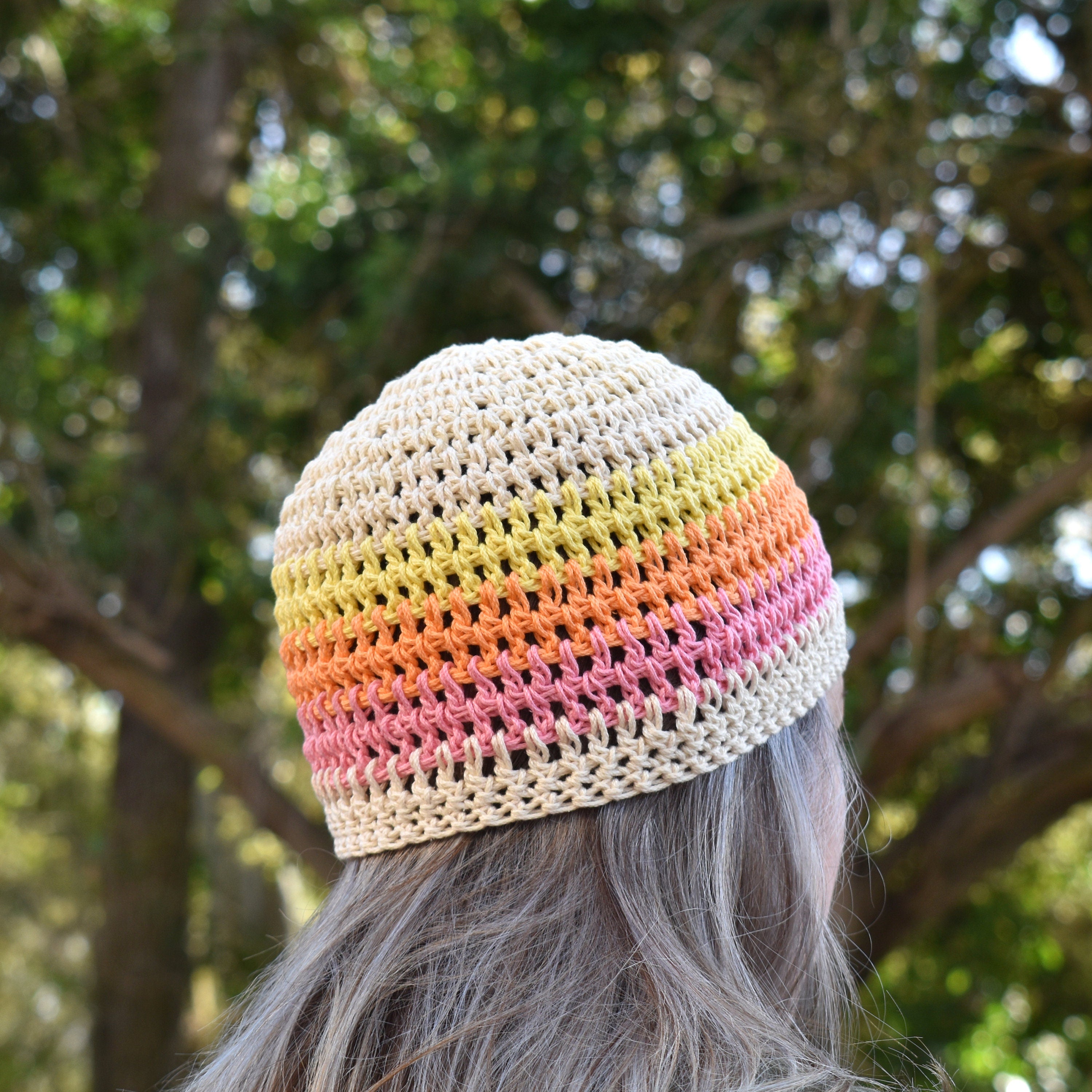 A person wearing a beige crochet beanie with yellow, orange, and pink stripes outdoors with trees in the background.