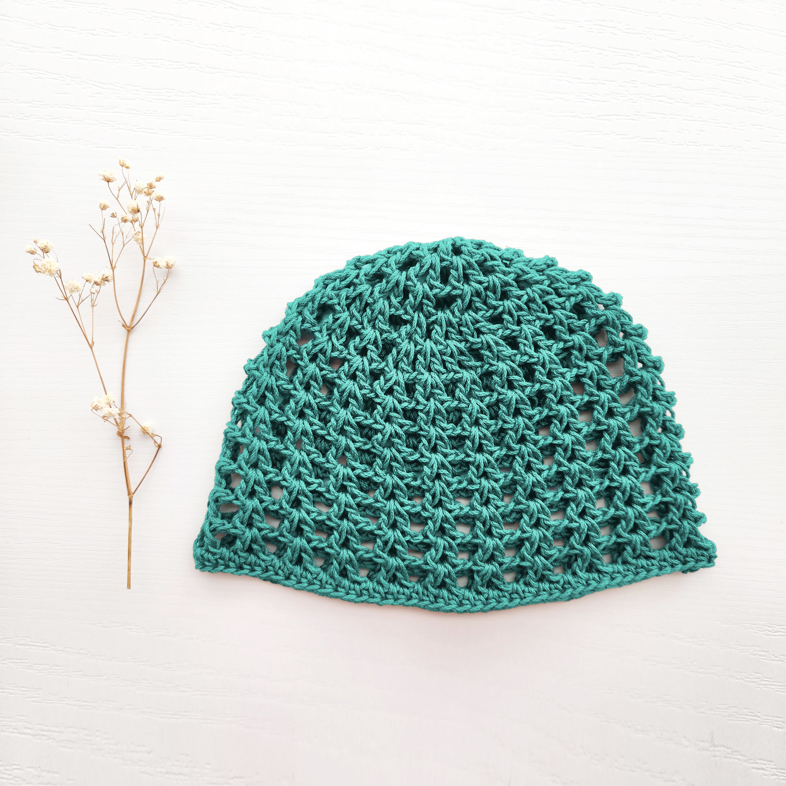 Light teal knitted mesh hat next to a sprig of flowers on a white background.