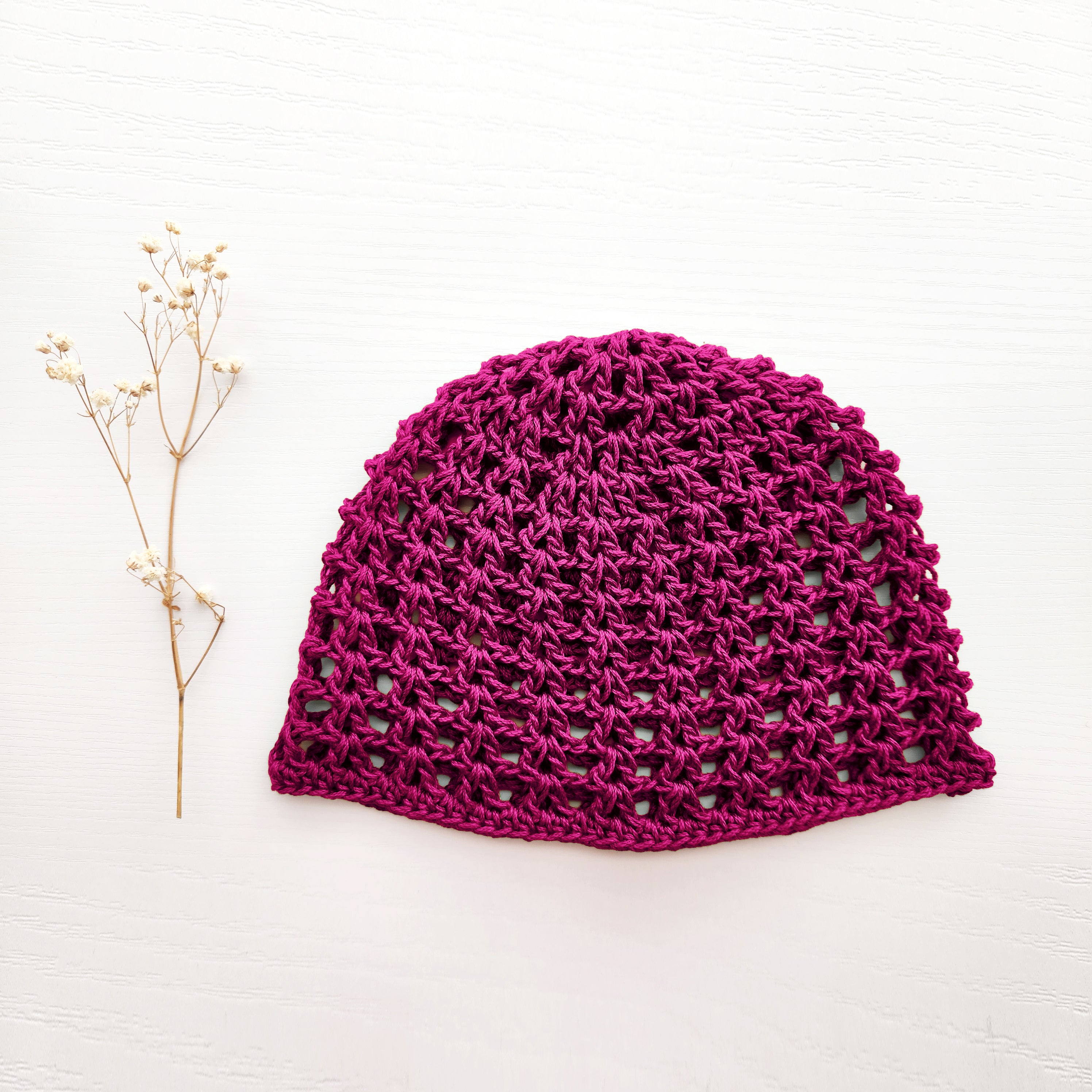 Bright magenta crochet hat on a white background with a sprig of dried flowers.