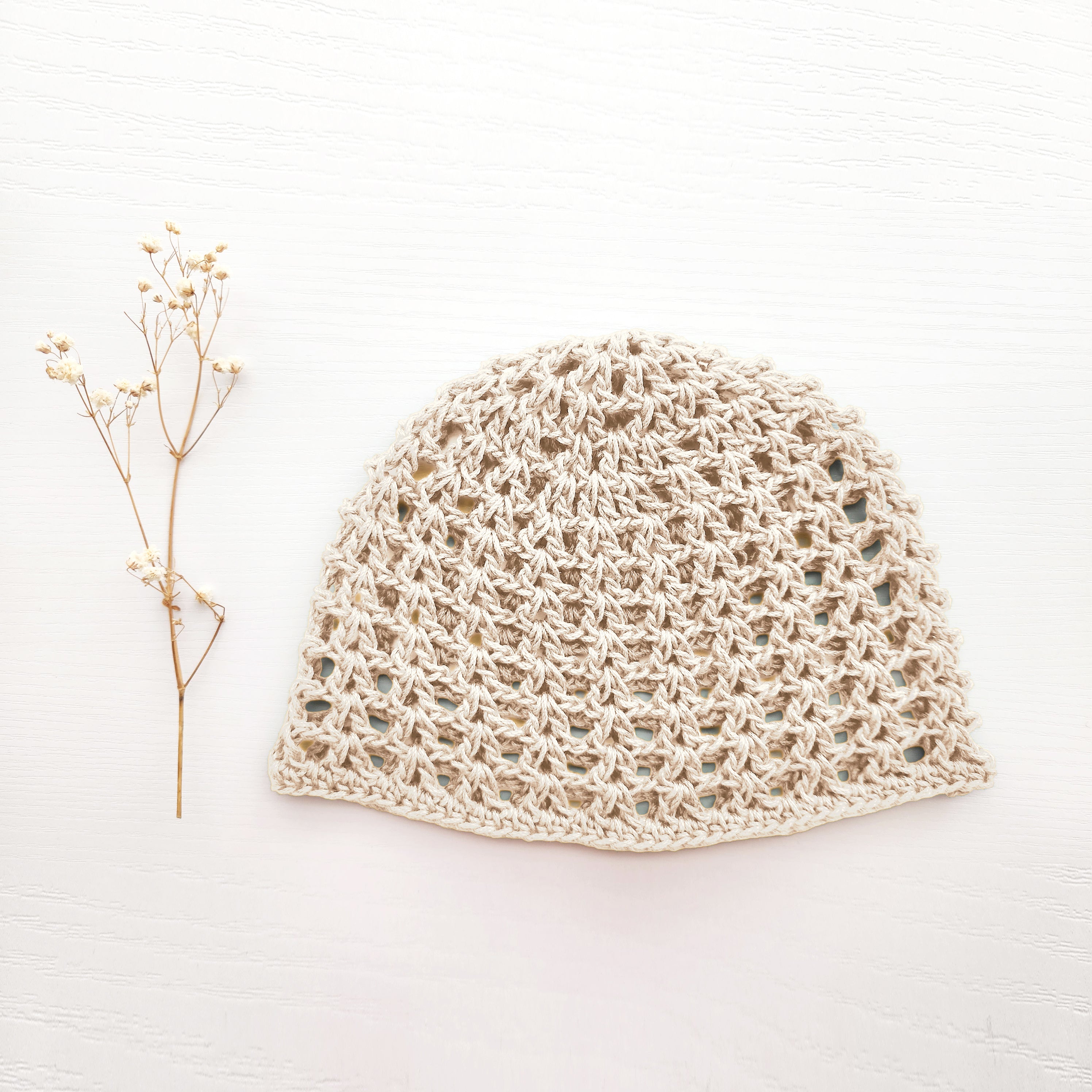 Beige crochet hat next to a sprig of dried flowers on a white background.