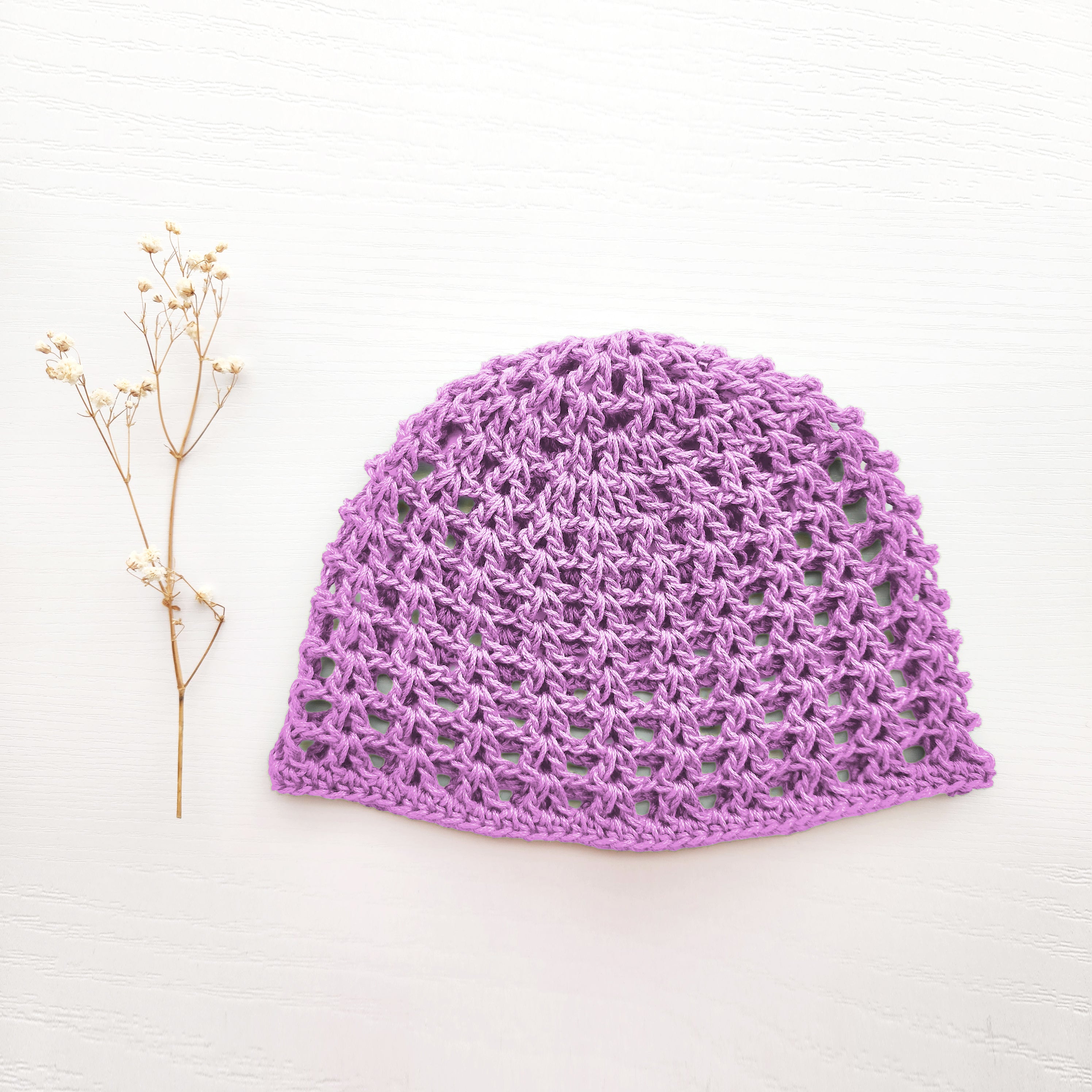Light purple crocheted hat on a white background with a sprig of dried flowers.