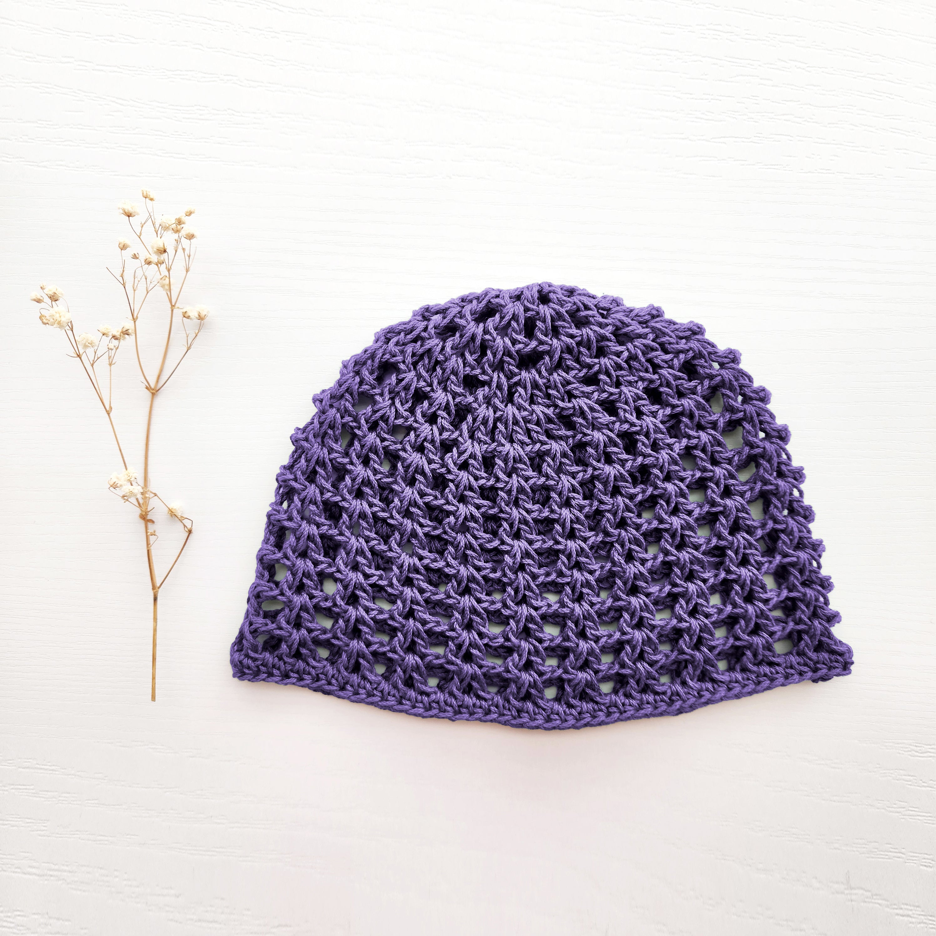 Purple knitted mesh summer cap next to a sprig of dried flowers on a white background.