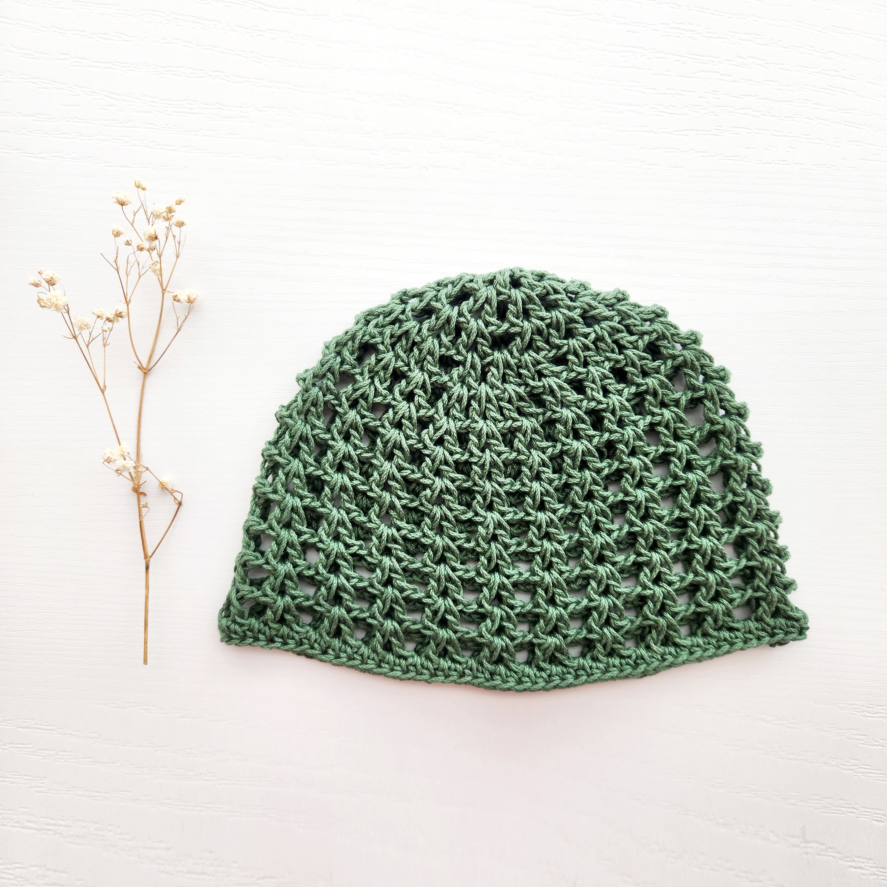 Green knitted mesh hat next to a sprig of flowers on a white background.