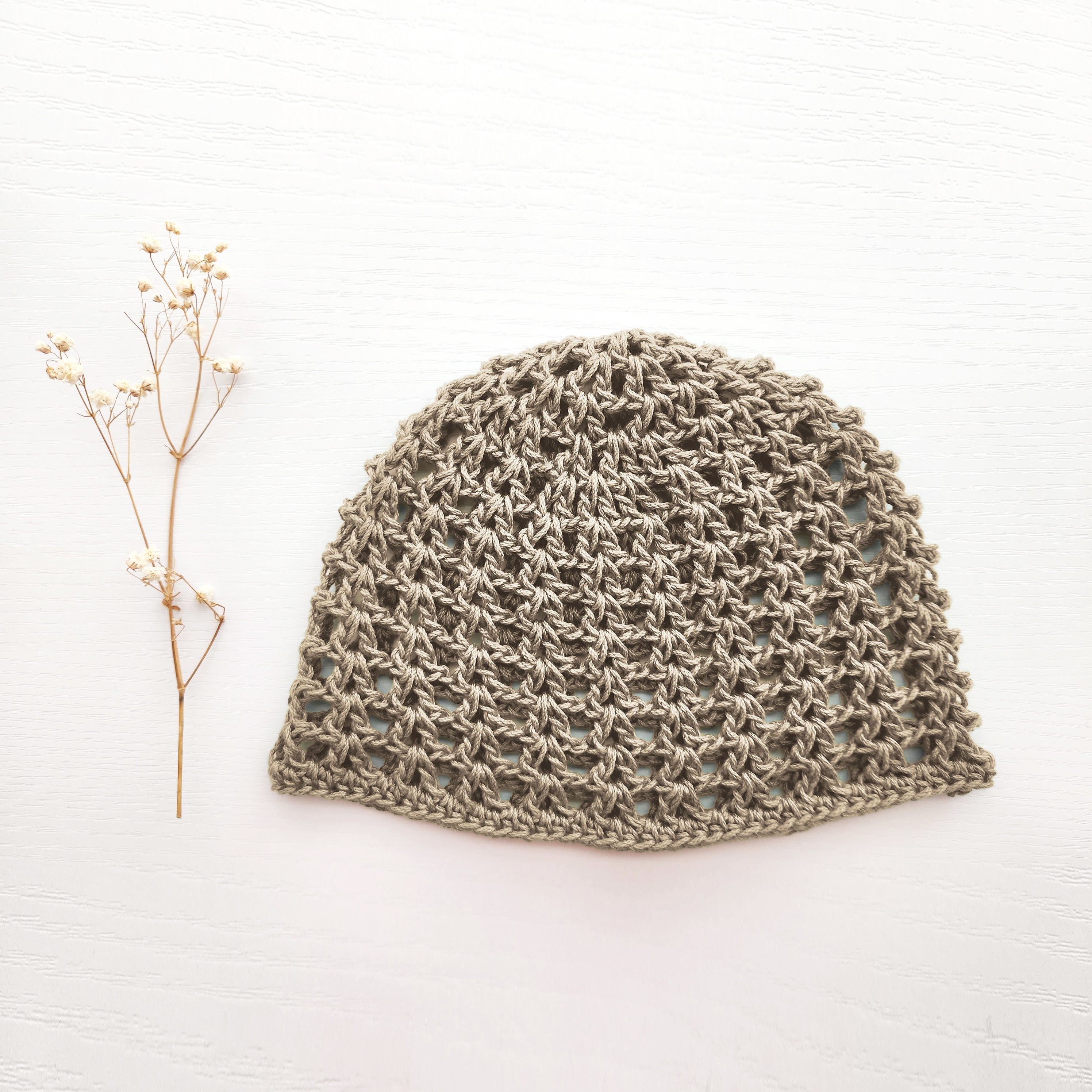 Taupe knitted mesh hat next to a branch of flowers on a white background.